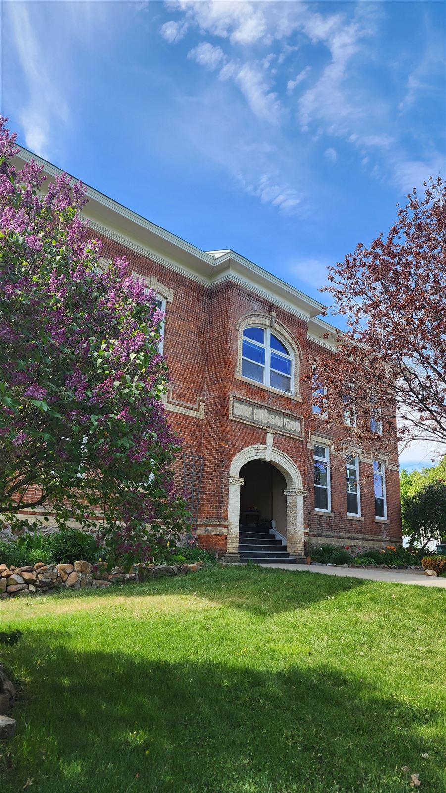 The front of the brick schoolhouse, with an arched doorway and a lilac bush in bloom.