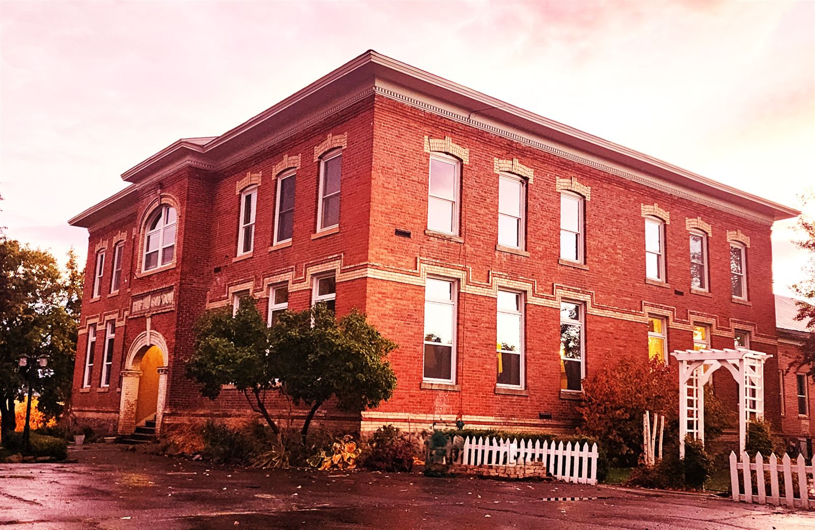 A two-story red brick schoolhouse with an arched entrance, white trim, and a white picket fence along the front.